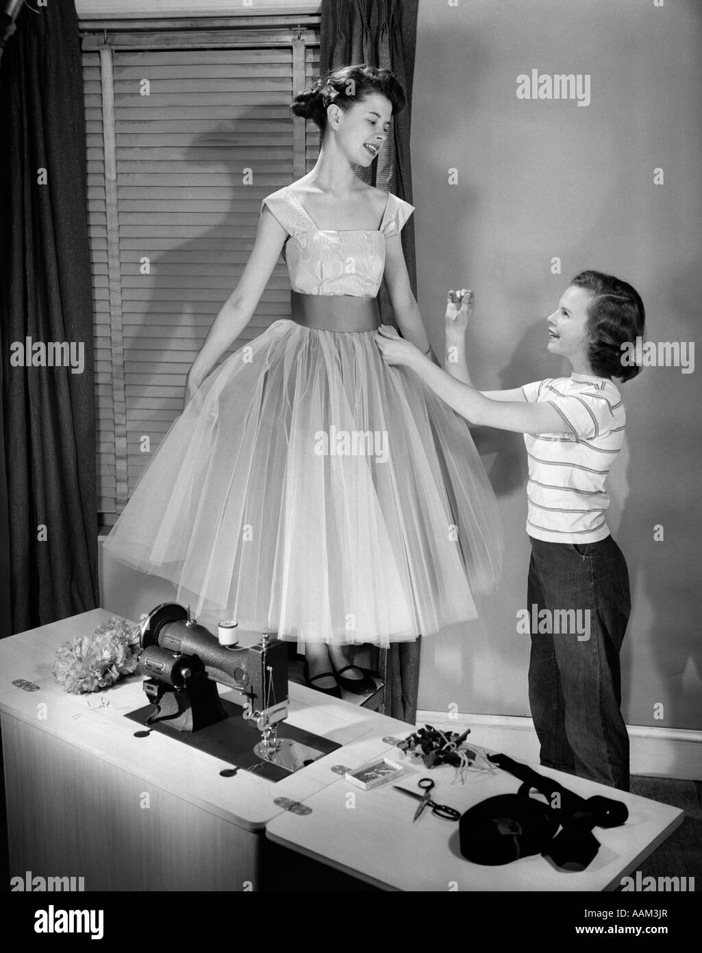 1950s TWO TEEN GIRLS WITH SEWING MACHINE ONE GIRL MODELING FANCY PROM DRESS OTHER GIRL ADJUSTING WAISTBAND Stock Photo