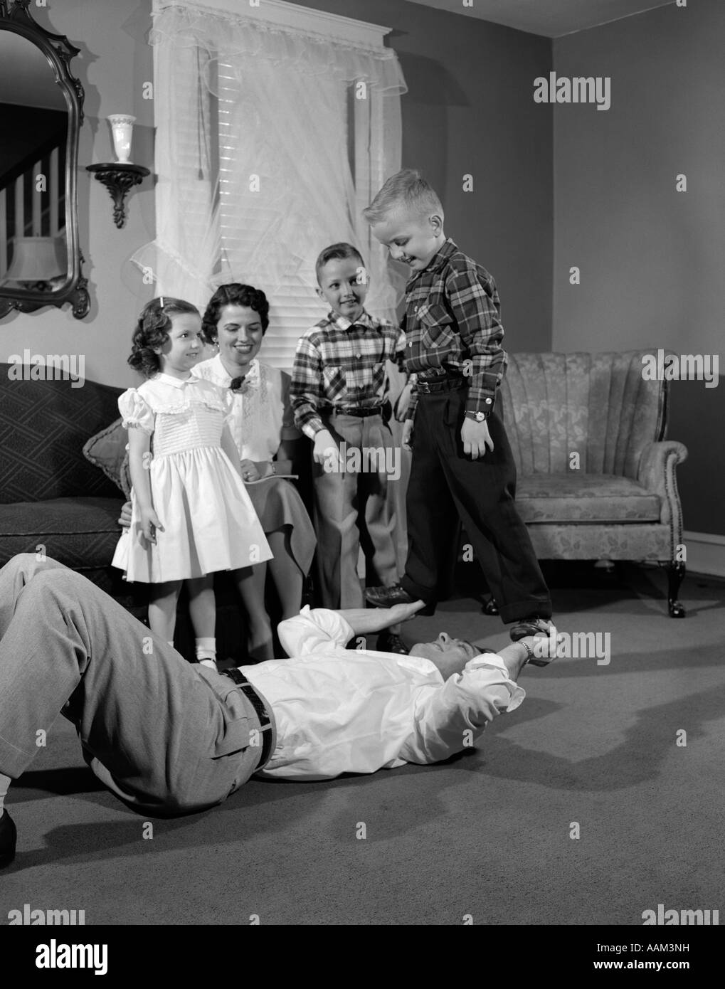 1950s FAMILY FATHER MOTHER DAUGHTER SONS PLAYING GAME FATHER LYING ON BACK LIFTING UP BOY STANDING ON HIS HANDS Stock Photo