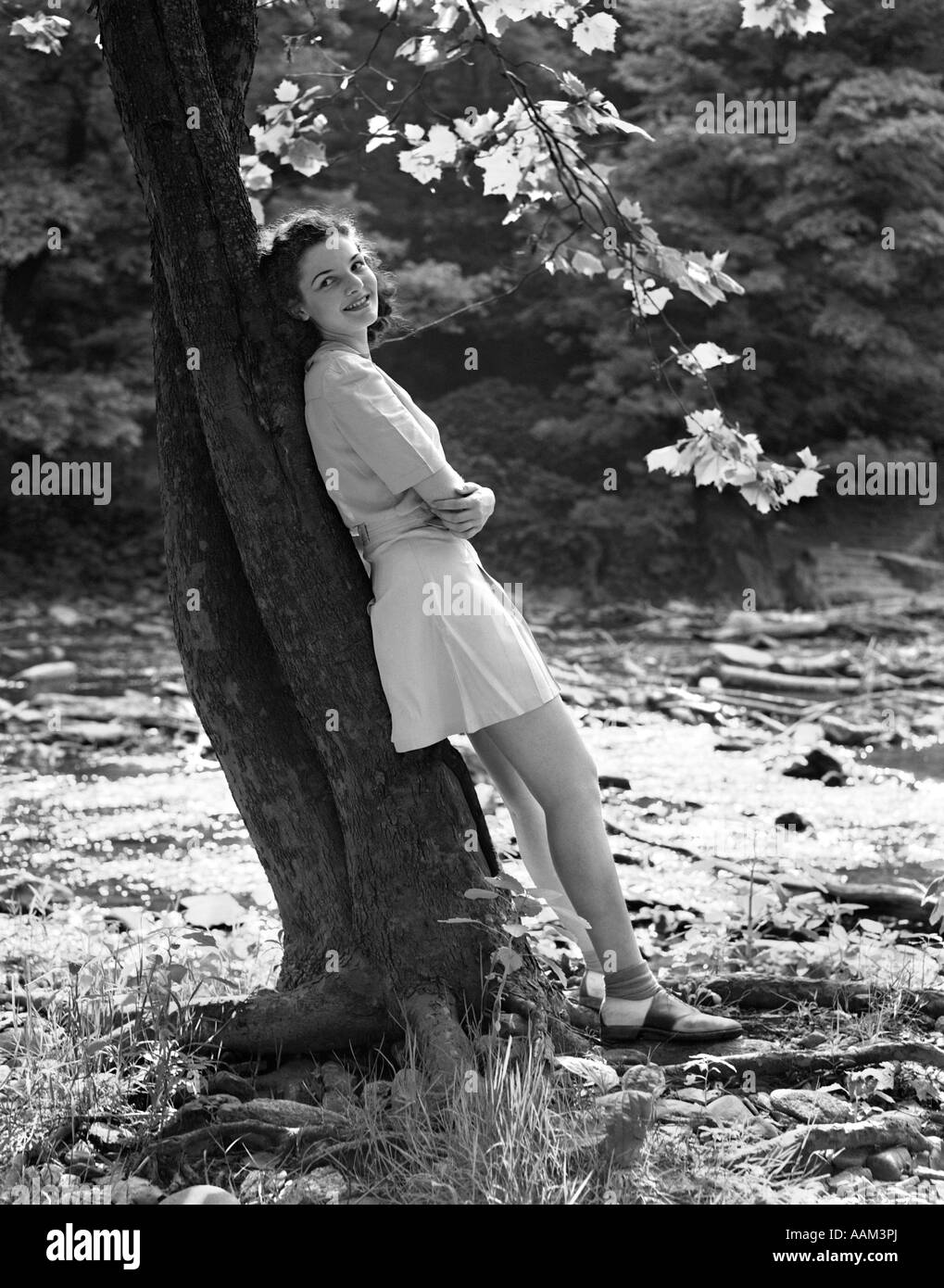 1940s SMILING YOUNG WOMAN WEARING SHORT SKIRT AND SADDLE SHOES LEANING AGAINST TREE BY STREAM Stock Photo