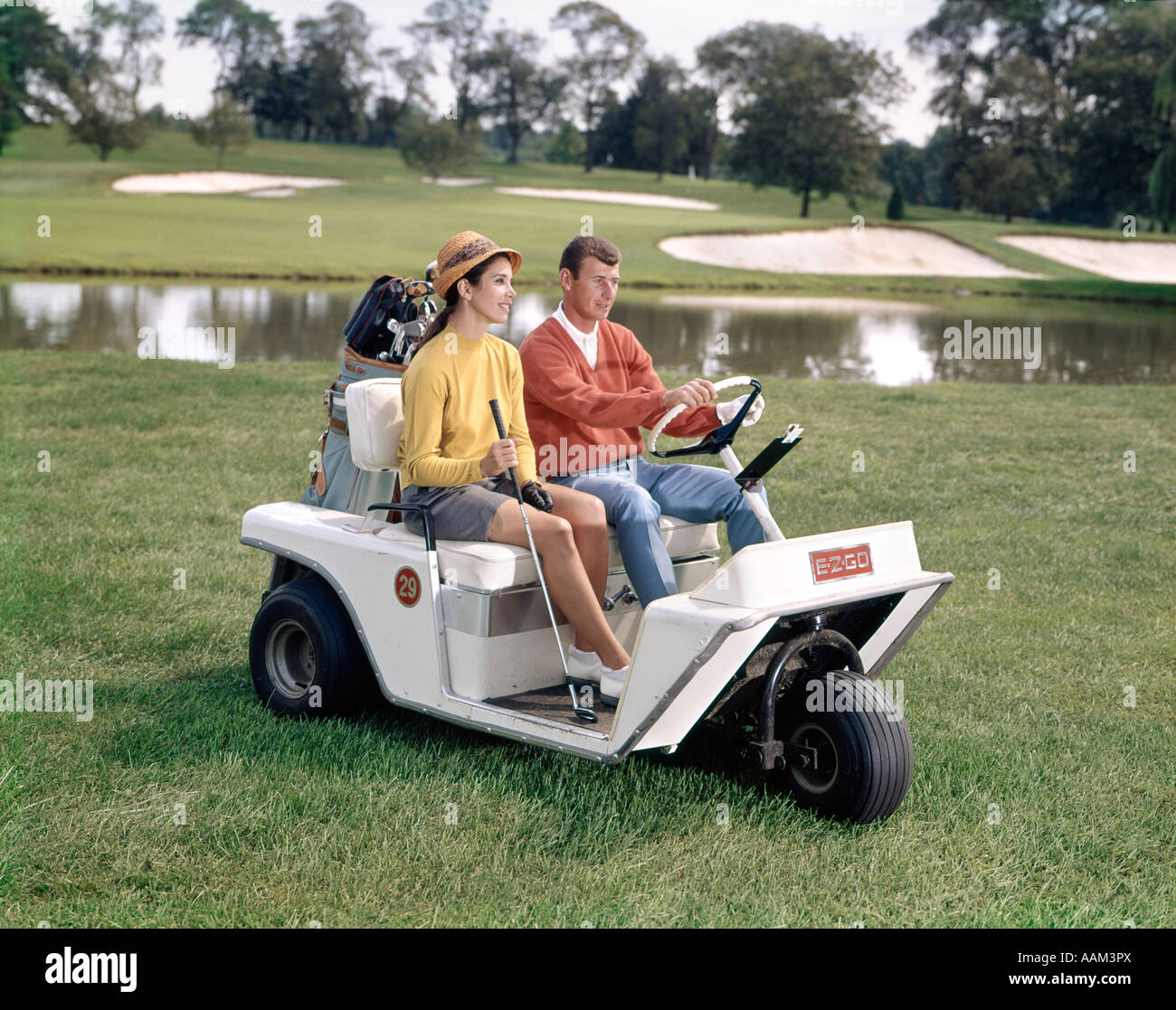1960s MAN AND WOMAN IN GOLF CART DRIVING ACROSS GRASS NEAR WATER COUPLE HOBBY LIFESTYLE GOLF CLUBS BAG Stock Photo