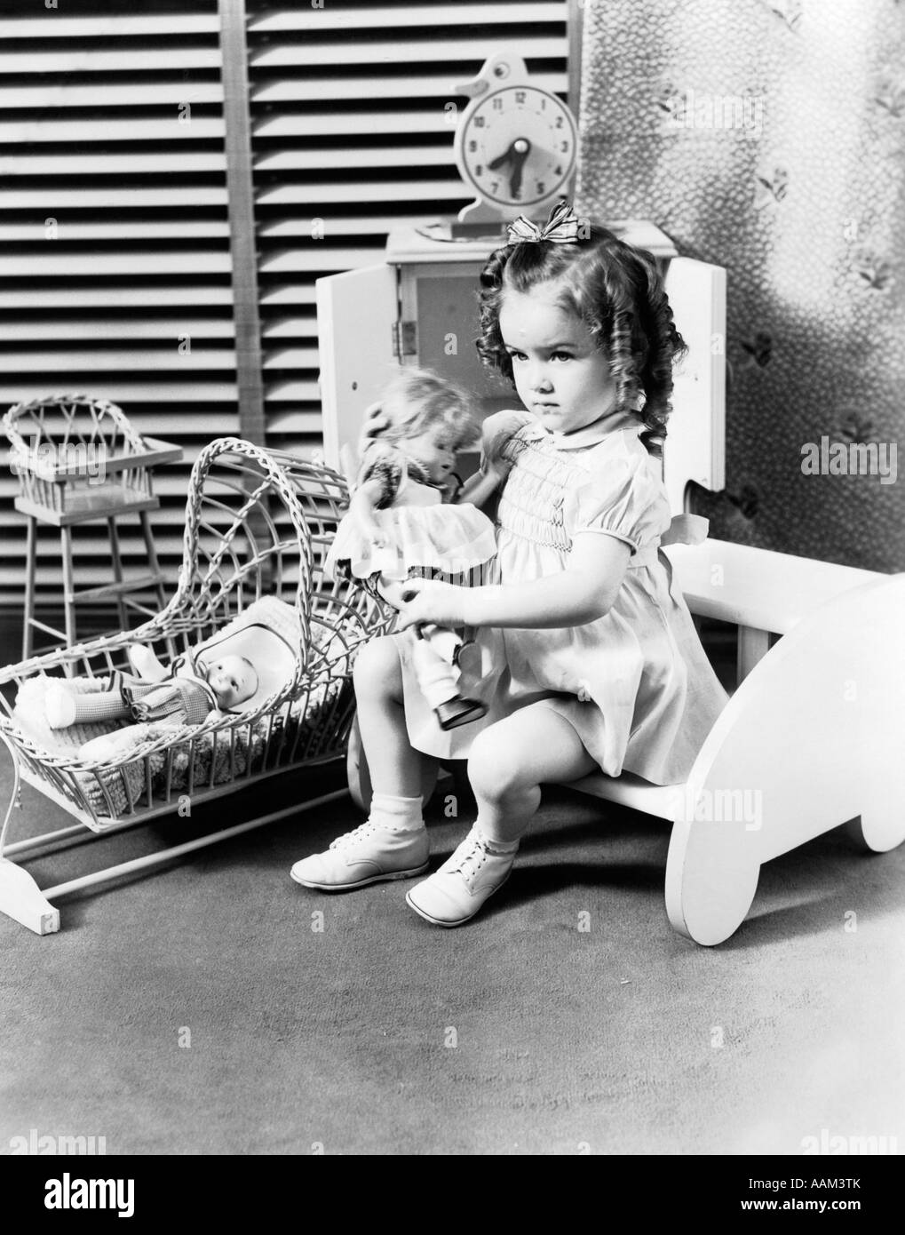 1940s 3 YEAR OLD GIRL SITTING HOLDING BABY DOLL TOY CRADLE TOY HIGH CHAIR & TOY CLOCK VENETIAN BLINDS Stock Photo