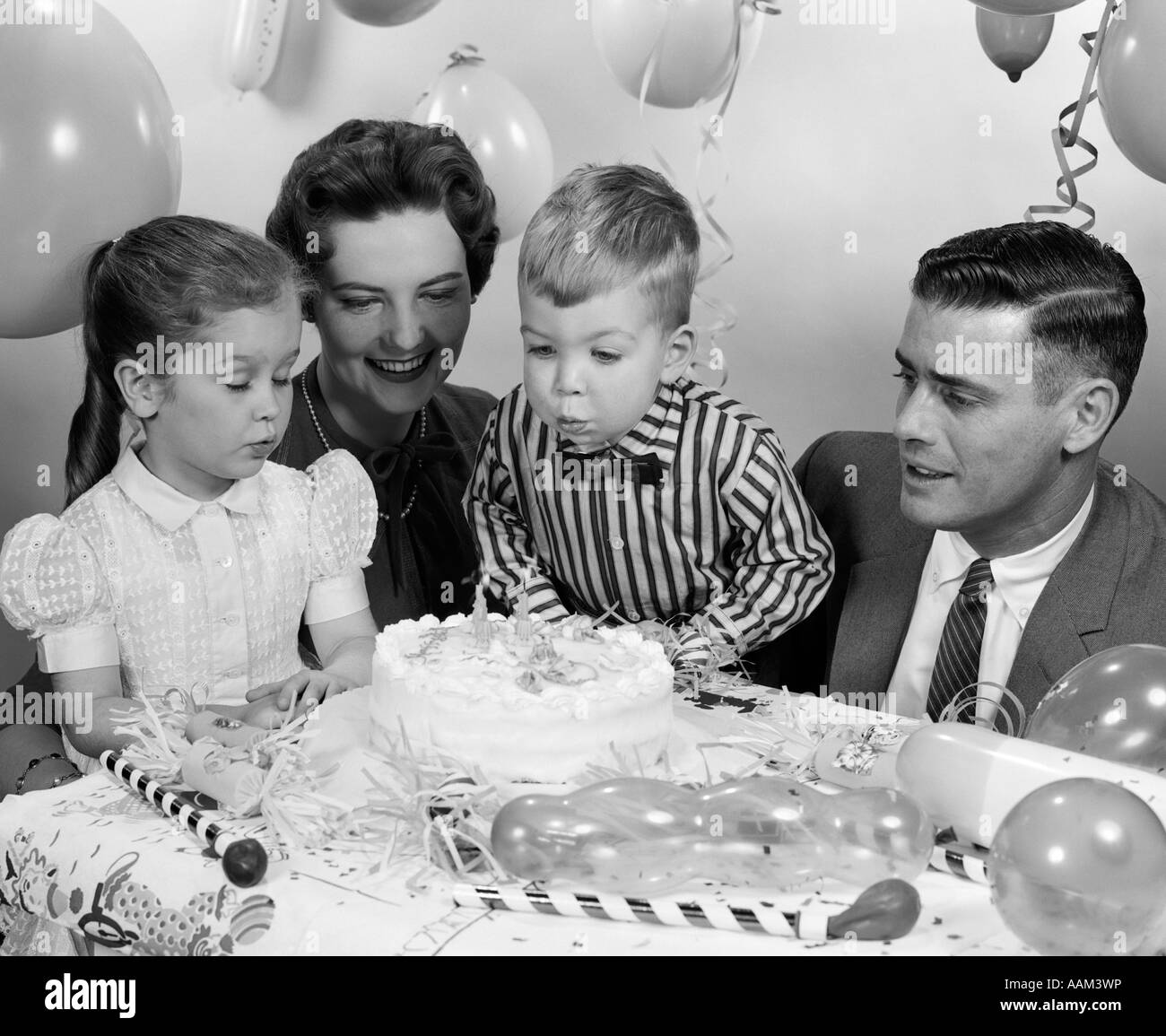 1950s CLOSE-UP OF FAMILY OF 4 WITH SON BLOWING OUT 2 CANDLES ON HIS BIRTHDAY CAKE Stock Photo