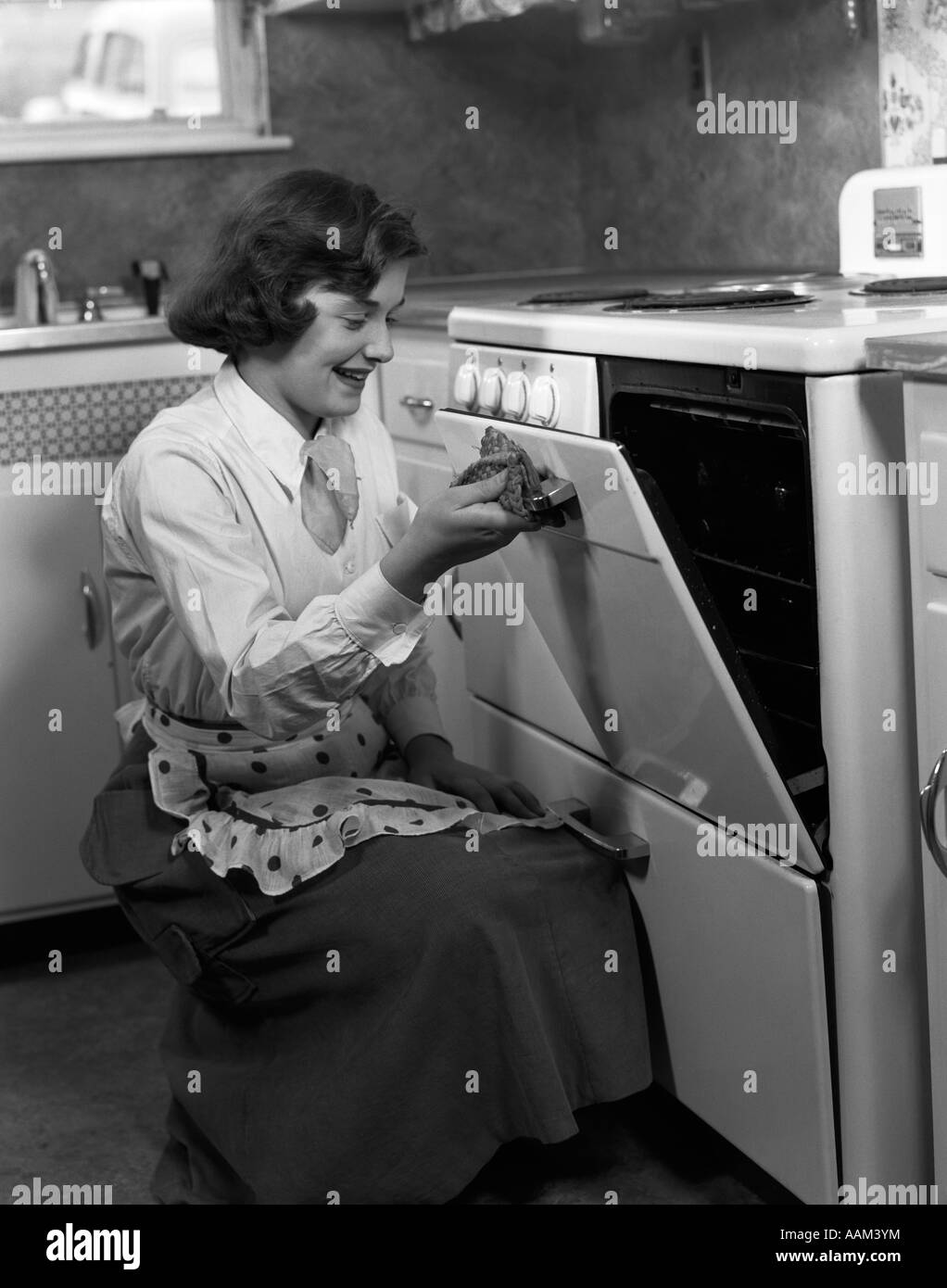 1950s YOUNG WOMAN GIRL WEARING APRON KNEELING OPENING OVEN DOOR KITCHEN STOVE COOKING Stock Photo