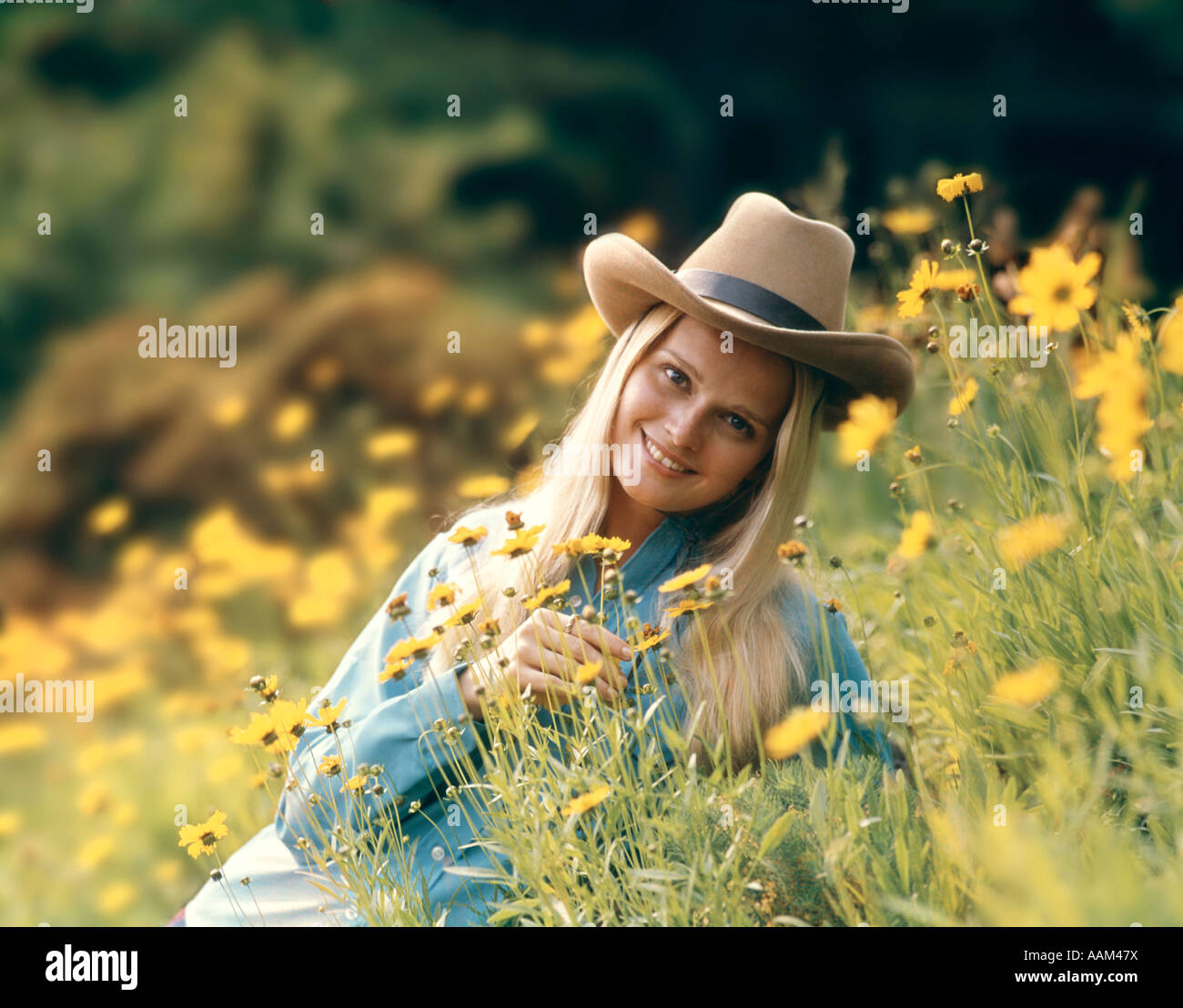 1970s RETRO BLOND WOMAN WEARING COWBOY HAT SITTING IN A FIELD OF BUTTERCUP WILD FLOWERS SMILING Stock Photo