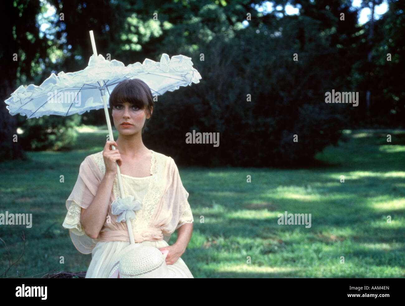 1970s WOMAN OUTDOOR PORTRAIT BROWN HAIR PINK WHITE DRESS WHITE HAT PINK RIBBON WHITE VICTORIAN PARASOL Stock Photo