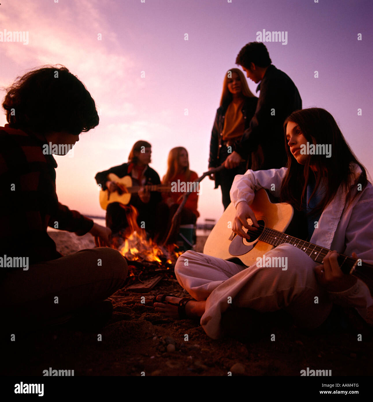 1970s TEENAGERS GATHERED AROUND BEACH CAMPFIRE PLAYING ACOUSTIC GUITARS Stock Photo