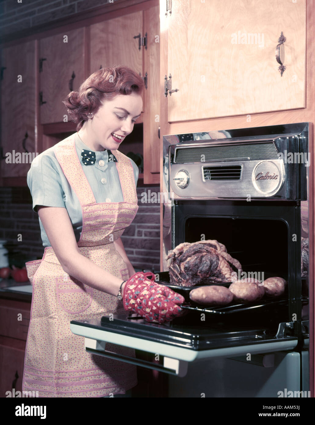 1950s SMILING WOMAN HOUSEWIFE WEARING APRON AND OVEN MITTS TAKING ROAST  BEEF WITH POTATOES OUT OF KITCHEN OVEN Stock Photo - Alamy
