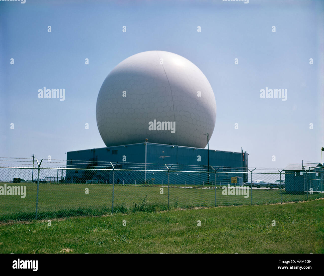 1970s EXTERIOR FACTORY PLANT INDUSTRIAL BUILDING SURROUNDED BY CHAIN LINK FENCE RADAR DOME SATELLITE RADIO COMMUNICATION Stock Photo