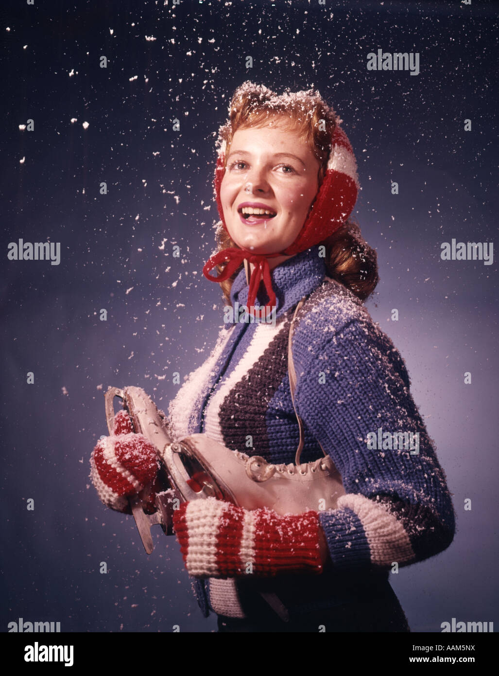 1960s SMILING YOUNG WOMAN WEARING RED MITTENS EAR MUFFS BLUE SWEATER HOLDING ICE SKATES IN FALLING SNOW Stock Photo