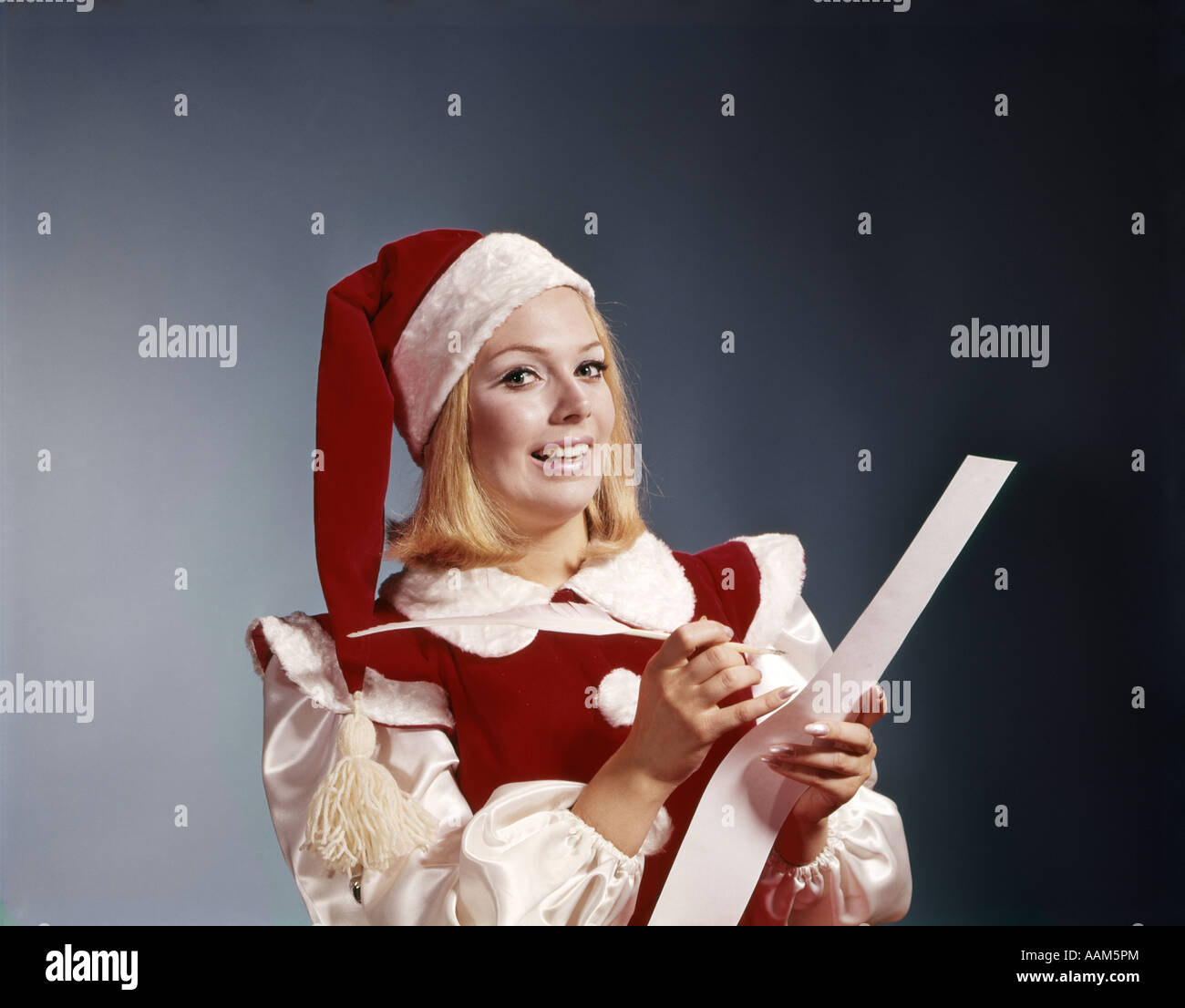 1960s YOUNG WOMAN IN SANTA HELPER HAT AND COSTUME HOLDING PEN AND LIST SYMBOLIC PORTRAIT Stock Photo