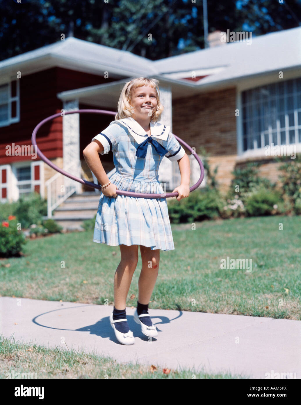 1950s 1960s LITTLE GIRL WITH HULA HOOP PLAYING OUTSIDE LAUGHING PURPLE ON SUBURBAN SIDEWALK IN SAILOR DRESS Stock Photo