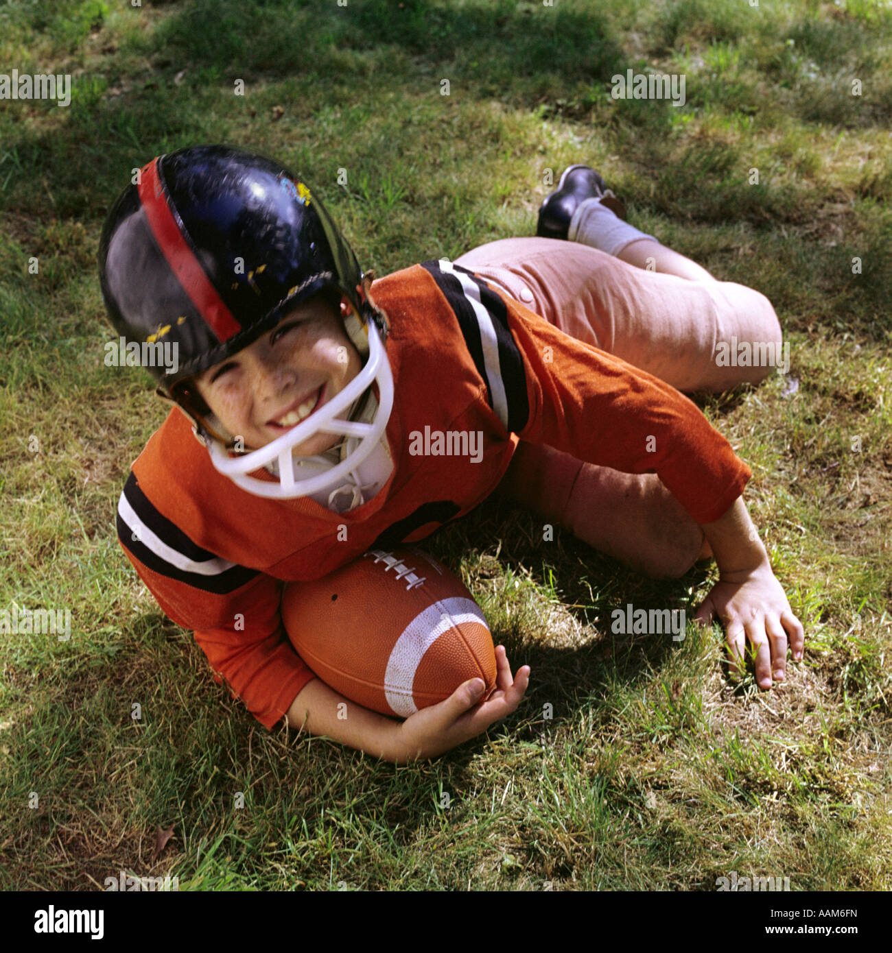 1970s FRECKLE FACED BOY FOOTBALL UNIFORM HELMET ORANGE TOP PINK PANTS LYING IN GRASS HOLDING FOOTBALL LOOK UP AT CAMERA Stock Photo