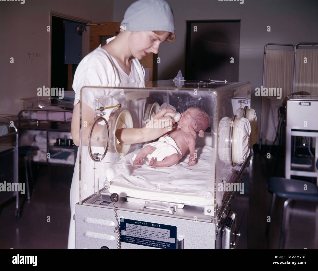 1960s WOMAN NURSE BOTTLE FEEDING INFANT BABY GIRL IN HOSPITAL NURSERY ISOLETTE PREMATURE INTENSIVE CARE Stock Photo