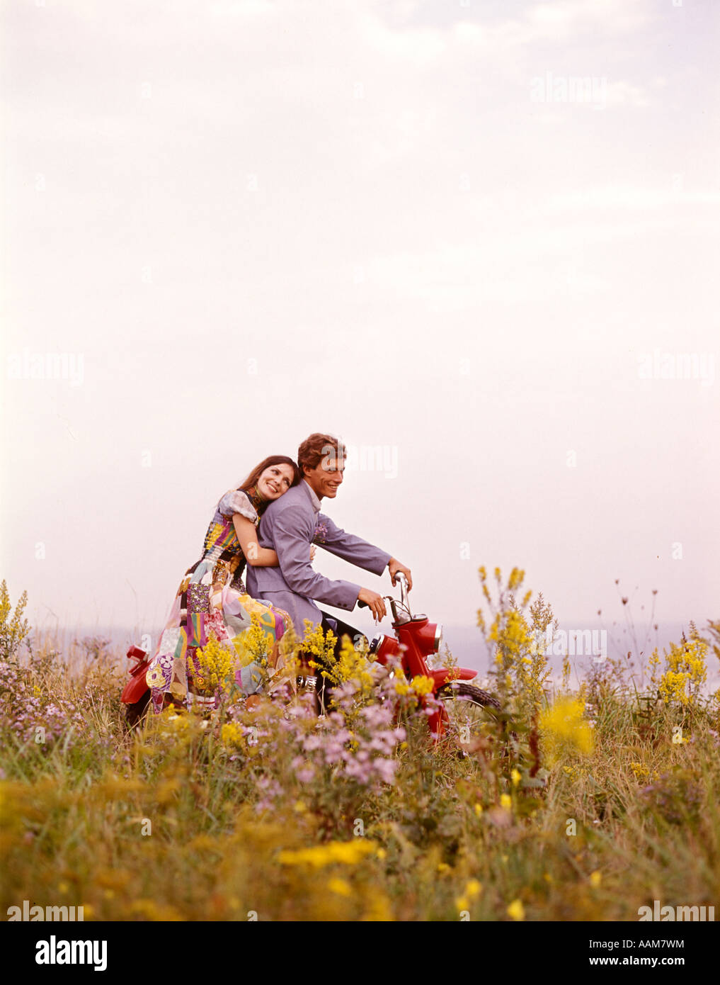 1970 1970s YOUNG COUPLE MAN WOMAN ROMANTIC RIDING RED MOTORCYCLE MOTOR BIKE FIELD FLOWERS Stock Photo