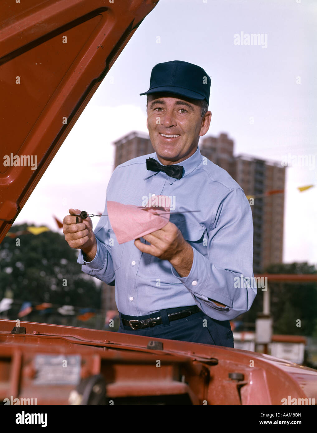 1960s MAN GAS SERVICE STATION MECHANIC CHECKING OIL DIP STICK UNDER CAR HOOD SMILING RETRO BLUE UNIFORM HAT BOW TIE Stock Photo