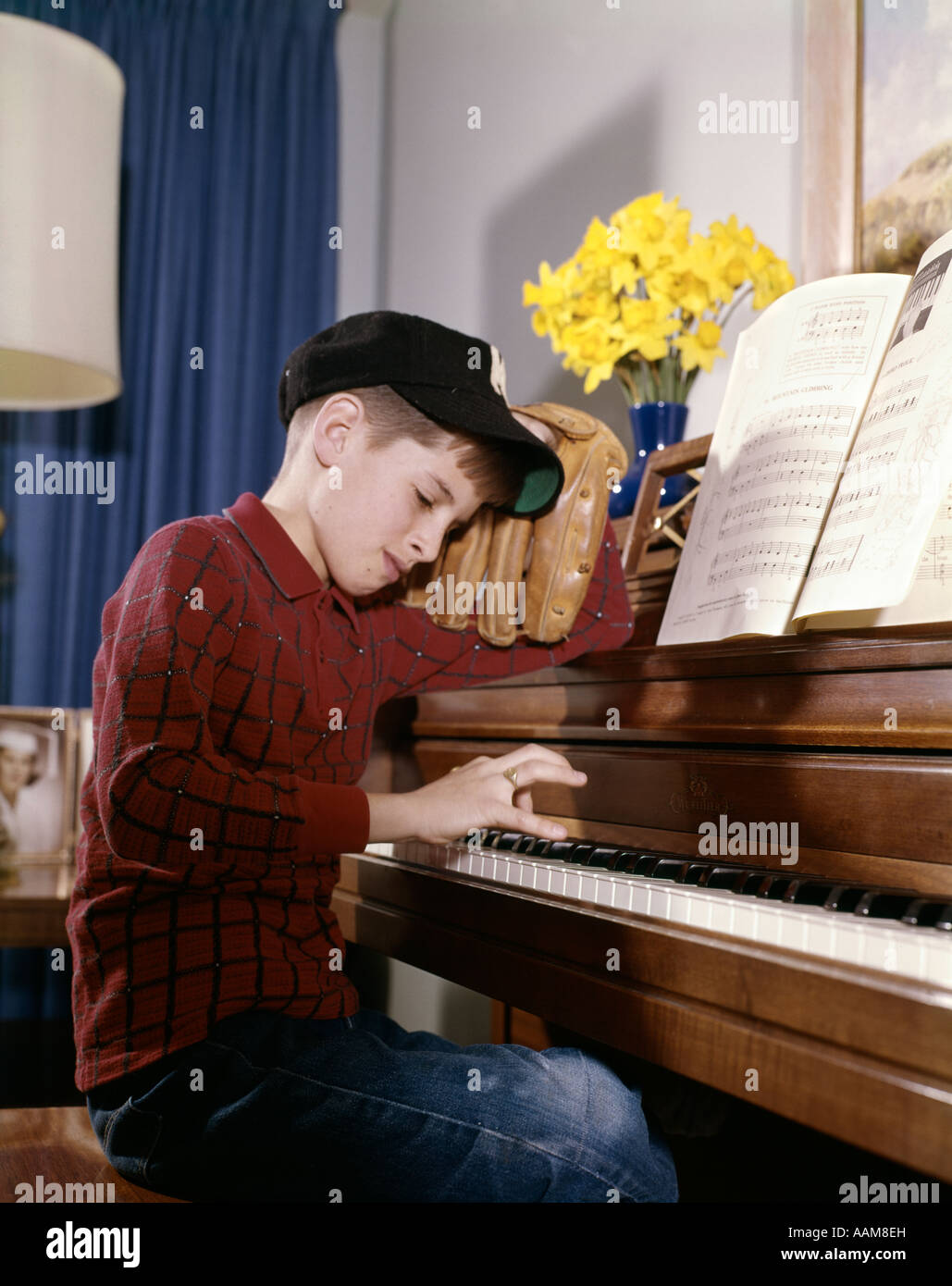 1960s ANNOYED LOOKING BOY WITH BASEBALL CAP AND GLOVE PRACTICING PIANO LESSON MITT IMPATIENT RESPONSIBILITY PRIORITY Stock Photo