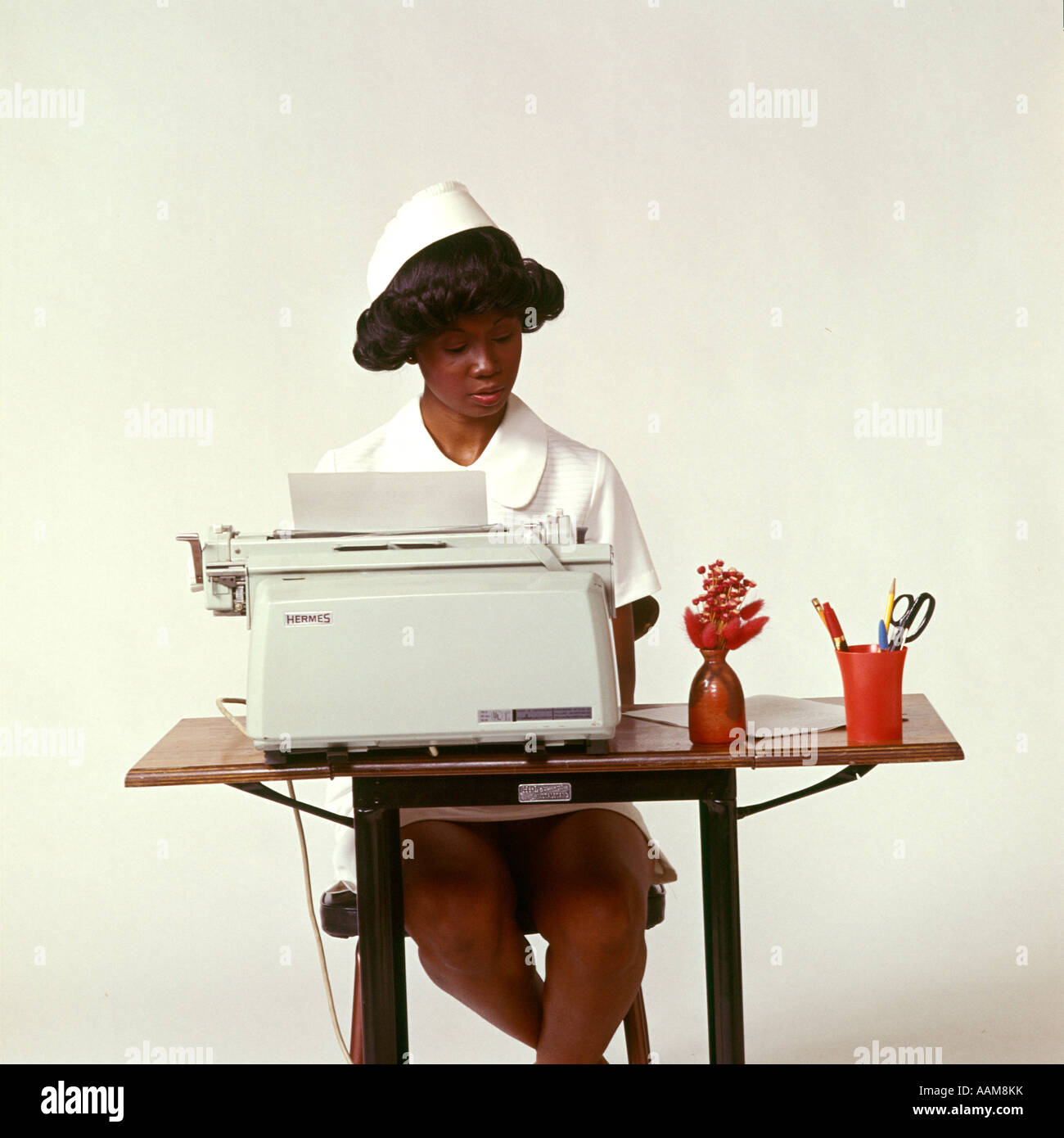 1970 1970s AFRICAN-AMERICAN WOMAN NURSE SEATED DESK TYPEWRITER TYPING DOCTORS OFFICE PAPERWORK NURSES MEDICAL HEALTH Stock Photo