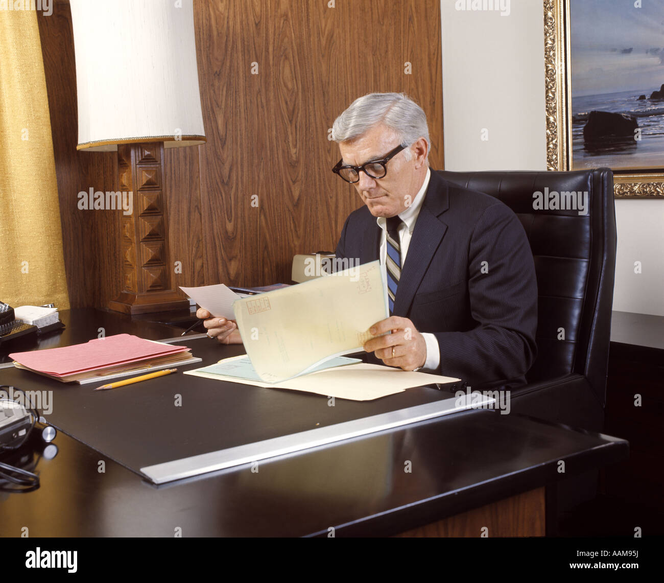 1960 1960s 1970 1970s MATURE MAN SILVER GRAY GREY HAIR EXECUTIVE SEATED DESK READING BUSINESS PAPERS MEN EXECUTIVES Stock Photo