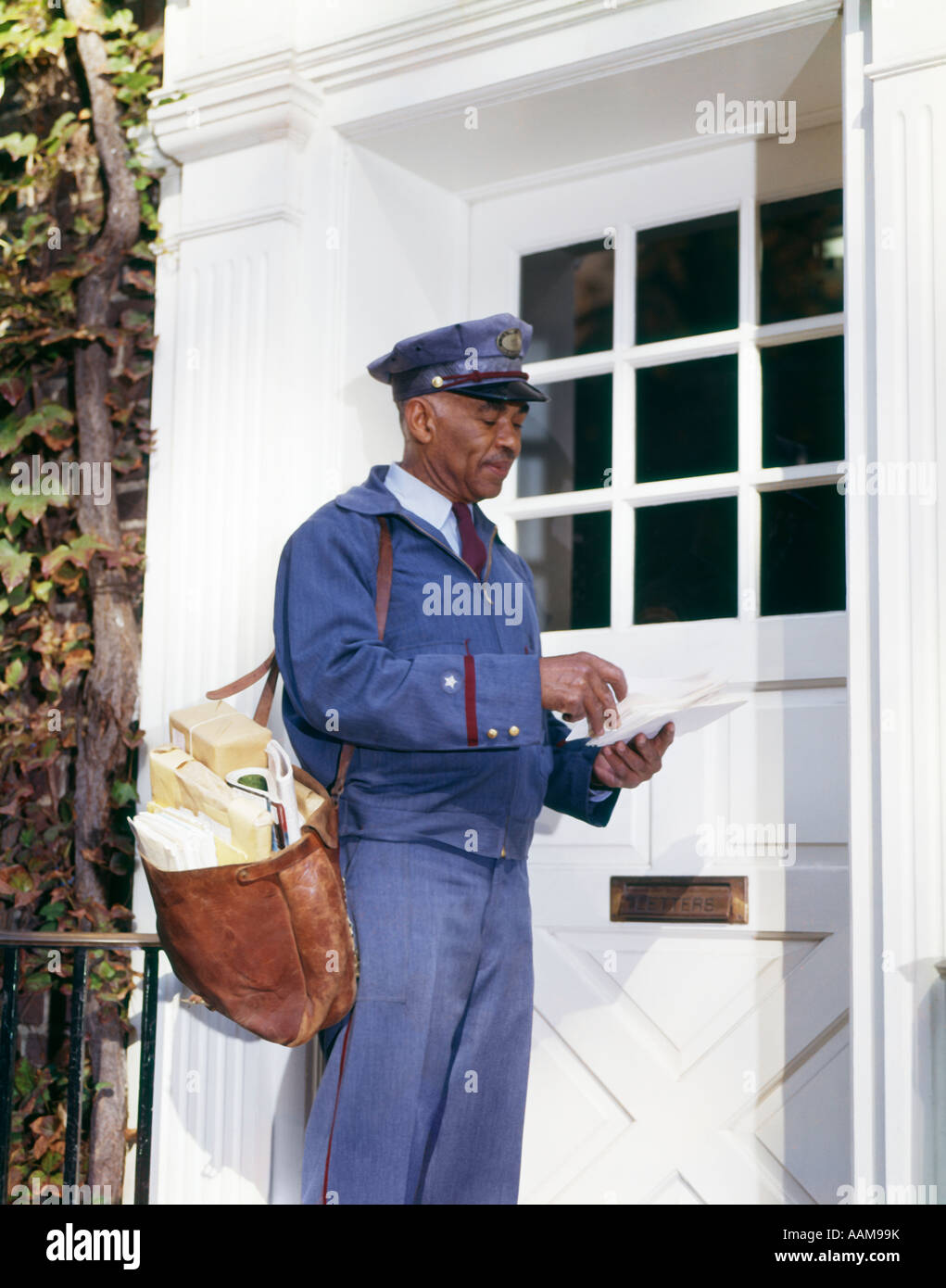 1960s AFRICAN-AMERICAN MAN MAILMAN RETRO CARRYING MAILBAG SORTING LETTERS FRONT DOOR DELIVERY DELIVERING USPS Stock Photo