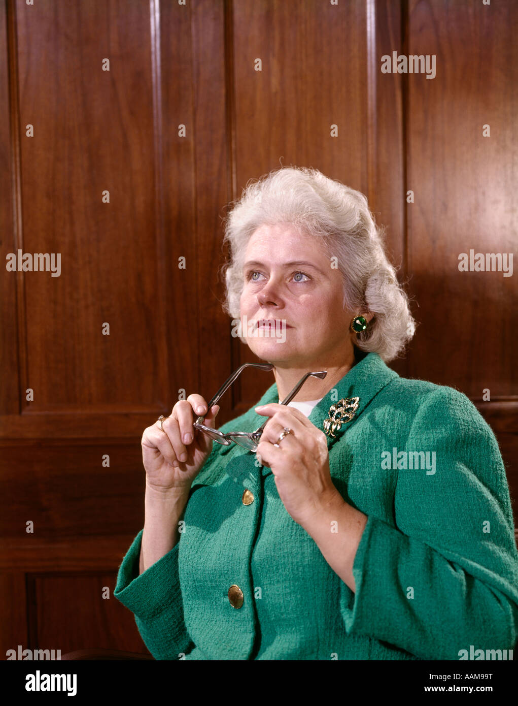 1960s SENIOR ELDERLY WOMAN THOUGHTFUL EXPRESSION SERIOUS HOLDING GLASSES IN HAND LOOK UP WOOD PANEL BACKGROUND Stock Photo