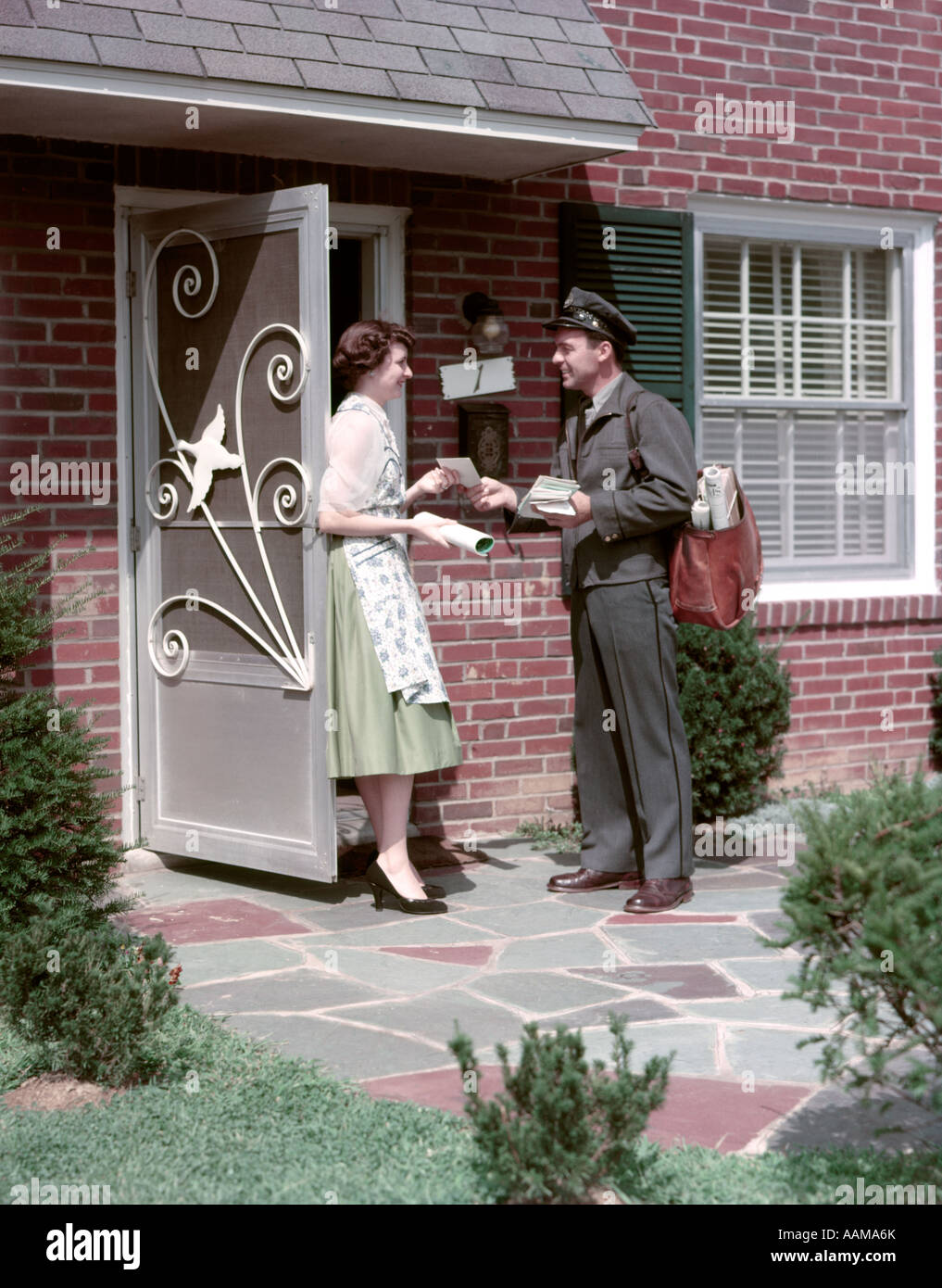 1950s MAILMAN DELIVERING MAIL TO WOMAN BRICK SUBURBAN HOME ALUMINUM SCREEN DOOR DELIVERY MAN HOUSEWIFE MAILBAG LETTER Stock Photo