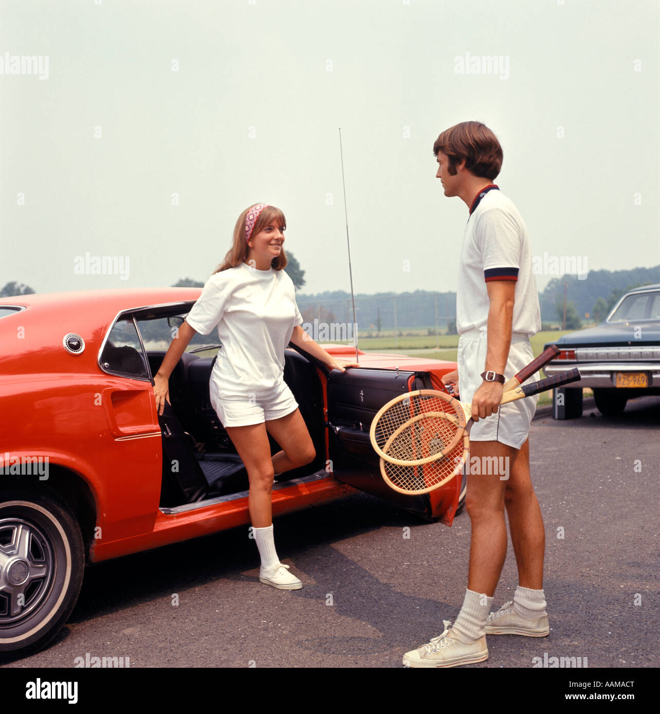 COUPLE GETTING OUT OF CAR MAN HOLDING TENNIS RACKETS RED WHITE 1970 1970s RETRO Stock Photo