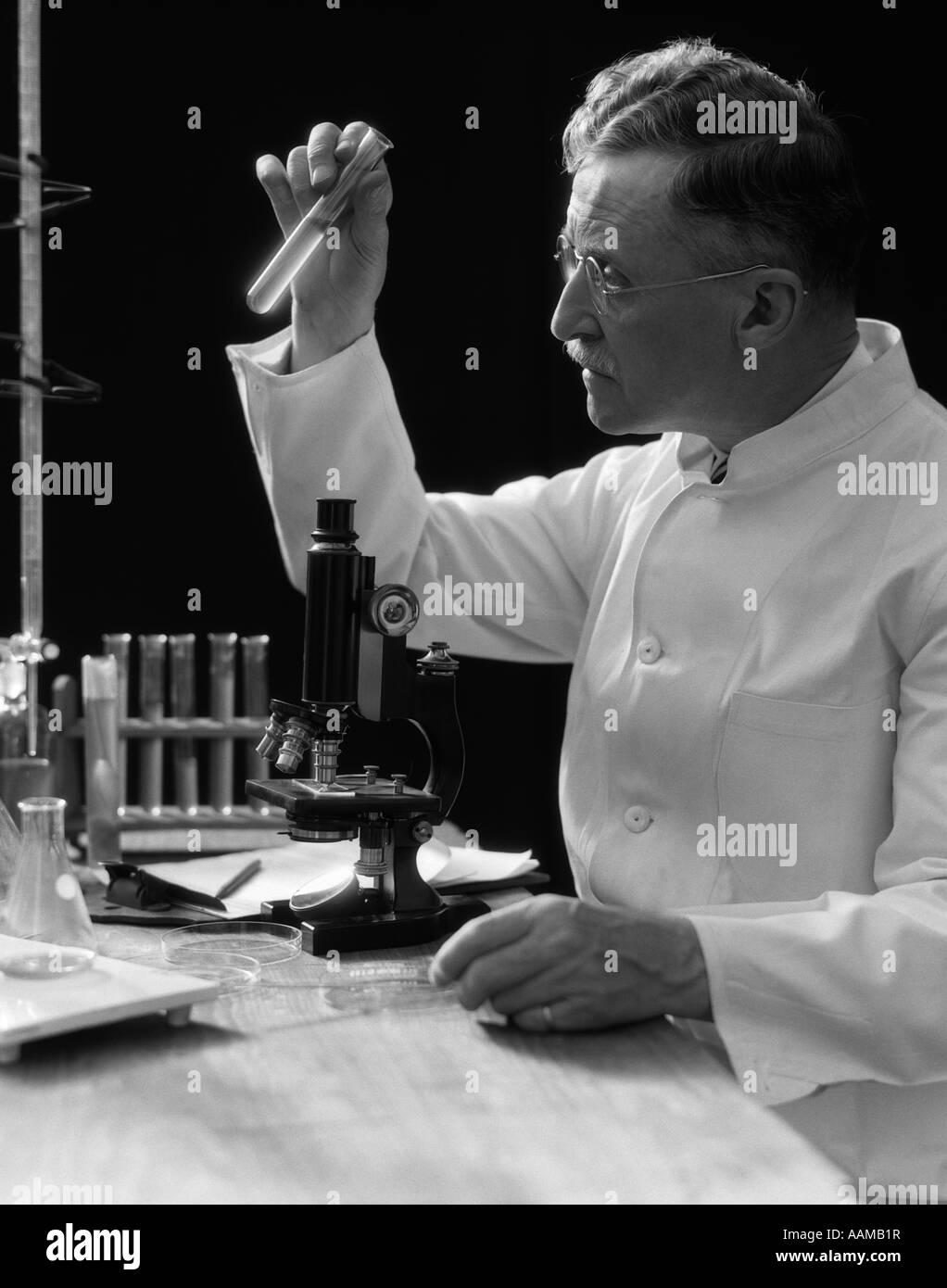 1920s 1930s 1940s SCIENTIST LAB TECHNICIAN IN WHITE COAT LOOKING AT TEST-TUBE IN FRONT OF MICROSCOPE Stock Photo