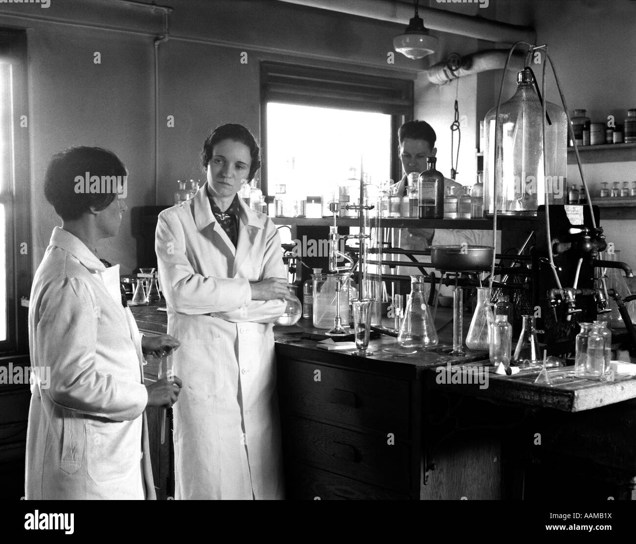 1930s TWO WOMEN ONE MAN WEARING LONG WHITE LAB COATS STANDING NEXT TO LAB TABLE CHEMICALS APPARATUS CONFERENCE BUNSEN BURNERS Stock Photo