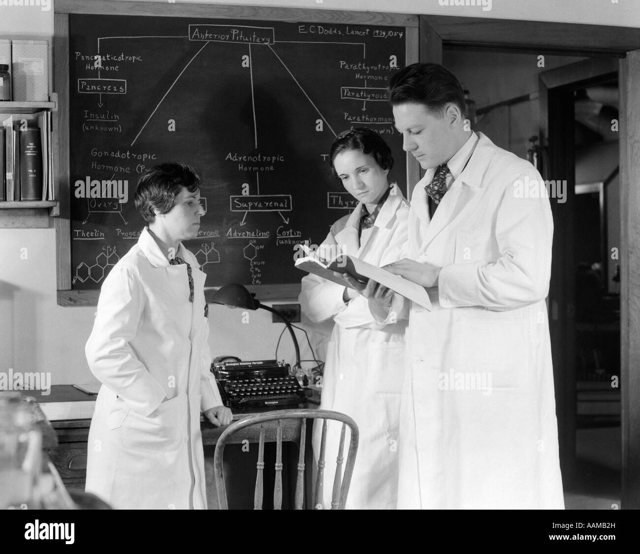 1930s 1940s THREE SCIENTISTS IN WHITE LAB COATS CONSULTING BOOK ONE MAN TWO WOMEN BLACKBOARD Stock Photo