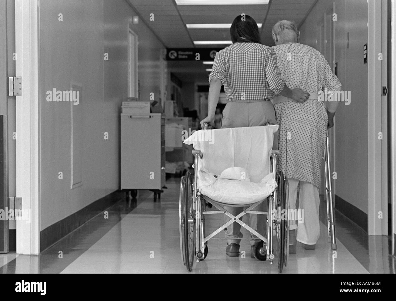 1980s REAR VIEW OF WOMAN WITH ARM AROUND ELDERLY MAN USING WALKER WHEELCHAIR BEHIND THEM Stock Photo