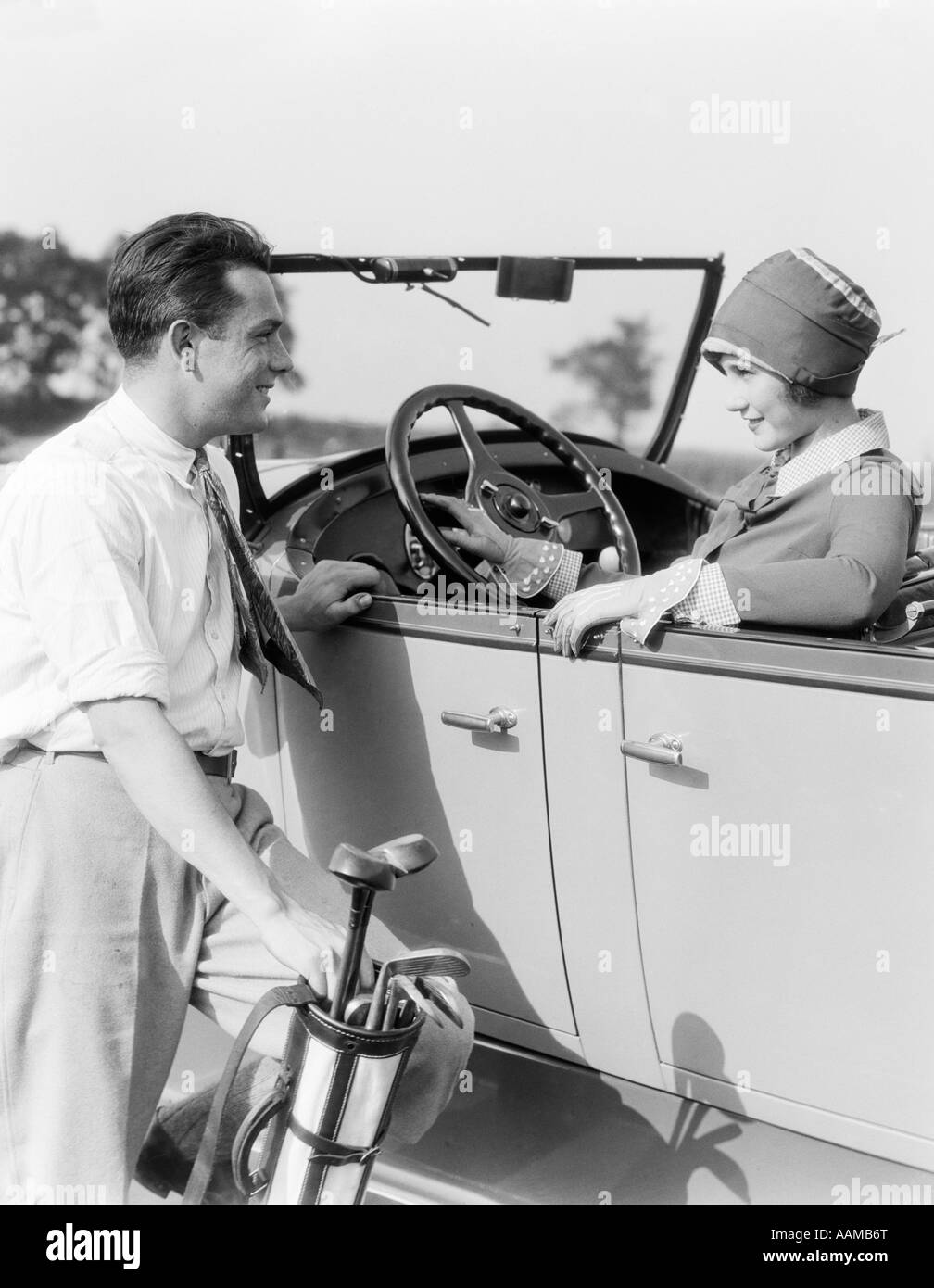 1920s 1930s MAN IN SHIRT AND TIE HOLDING GOLF BAG STANDING NEXT TO WOMAN SITTING IN A CONVERTIBLE SEDAN HAND ON STEERING WHEEL Stock Photo