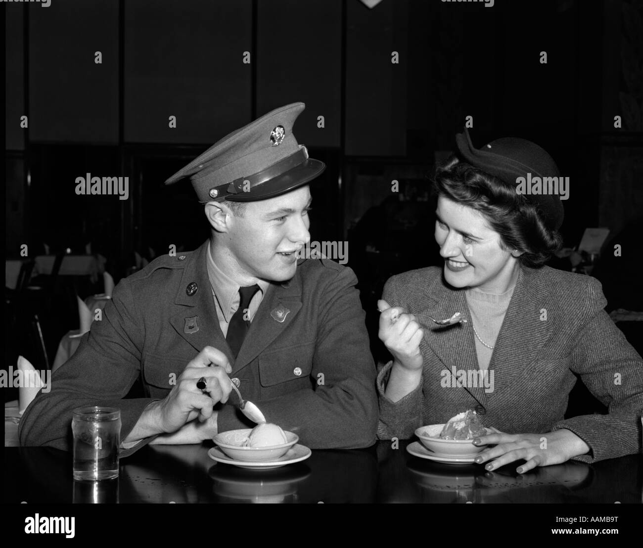 1940s SMILING COUPLE MAN SOLDIER IN ARMY UNIFORM AND WOMAN GIRLFRIEND SITTING AT SODA FOUNTAIN COUNTER EATING DISH OF ICE CREAM Stock Photo