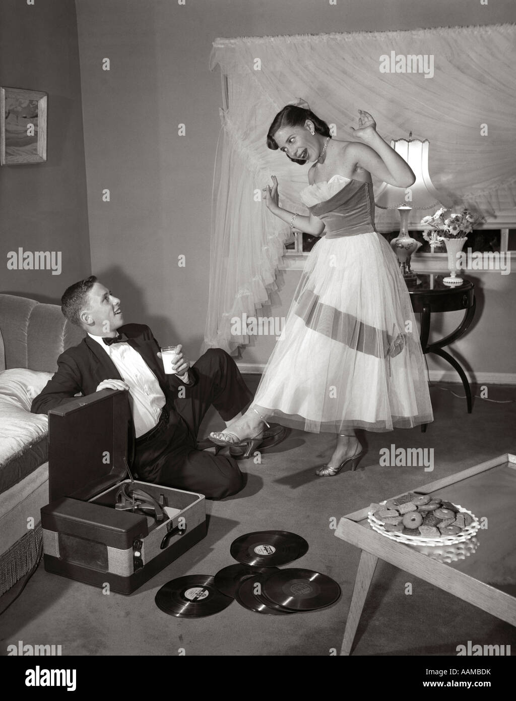 1950s 1960s TEEN COUPLE IN LIVING ROOM IN PROM DRESS & TUXEDO GUY SITTING ON FLOOR & GIRL DANCING TO MUSIC FROM A RECORD PLAYER Stock Photo