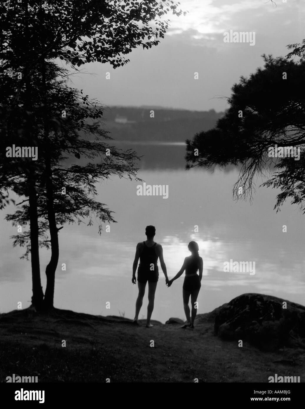 1930s REAR VIEW SILHOUETTE OF MAN AND WOMAN IN BATHING SUITS HOLDING HANDS WATCHING SUNSET LAKESIDE Stock Photo