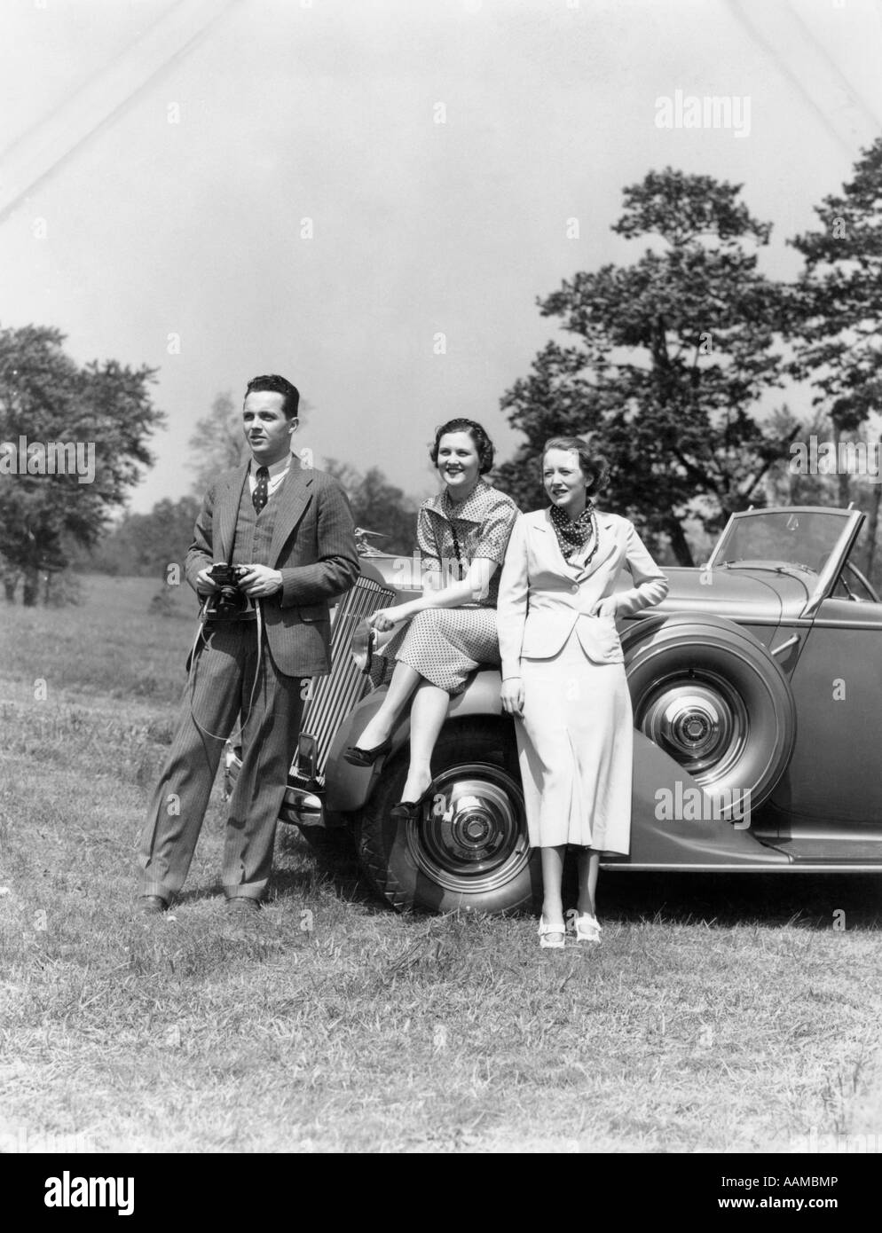 1930s MAN WITH CAMERA AND TWO WOMEN IN FRONT OF CONVERTIBLE CAR STYLE FASHION WEALTH SPECTATORS Stock Photo