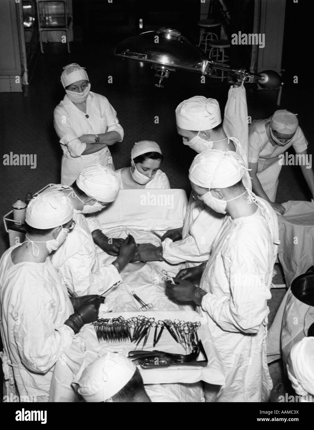 1930s SURGERY TEAM WEARING WHITE GOWNS CAPS AND MASKS IN HOSPITAL OPERATING ROOM PERFORMING A MEDICAL PROCEDURE Stock Photo