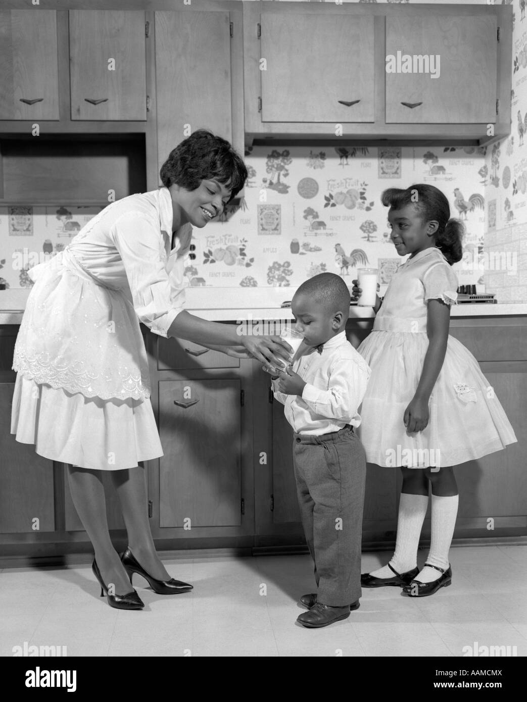 1960s SMILING AFRICAN AMERICAN WOMAN MOTHER IN APRON AND PUMPS GIVING A GLASS OF MILK TO SON AND DAUGHTER WITH MARY JANE SHOES Stock Photo