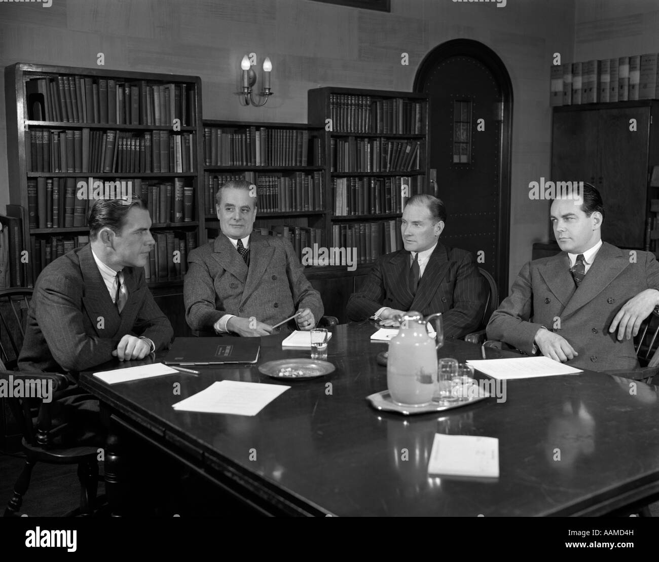 1940s GROUP FOUR BUSINESSMEN CORPORATE EXECUTIVES SIT AROUND CONFERENCE TABLE SERIOUS EXPRESSIONS MEETING DECISION PAPERS Stock Photo