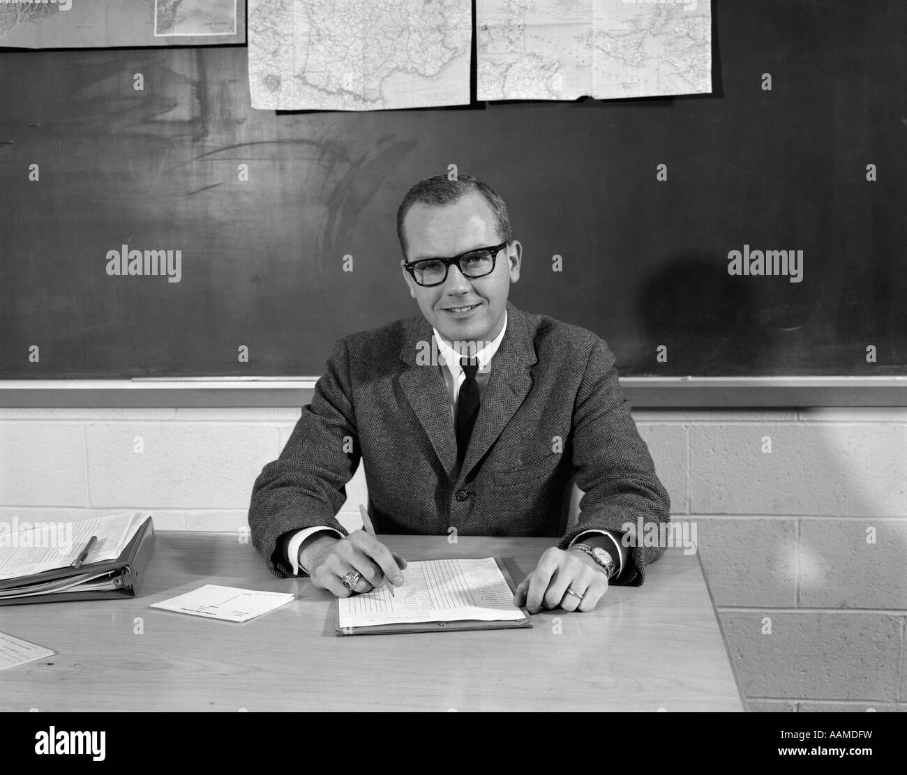 1960s MAN TEACHER AT DESK IN CLASSROOM PENCIL PAPER IN FRONT BLACKBOARD BACKGROUND SMILING AT CAMERA Stock Photo