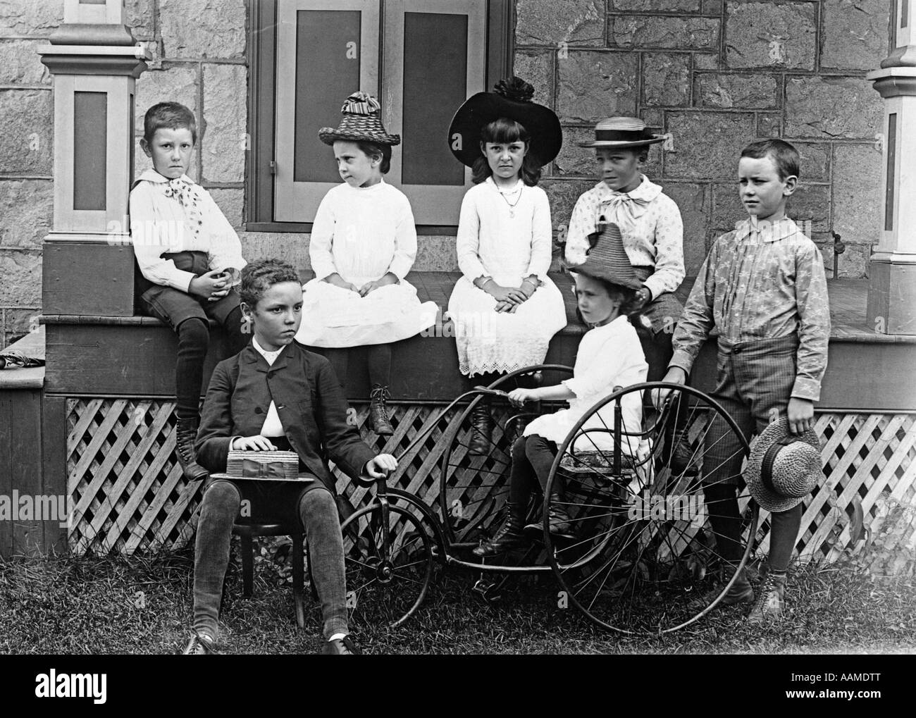 1890s 1900s TURN OF THE CENTURY GROUP OF SEVEN CHILDREN SITTING ON & AROUND PORCH ONE GIRL ON OLD FASHIONED TRICYCLE Stock Photo