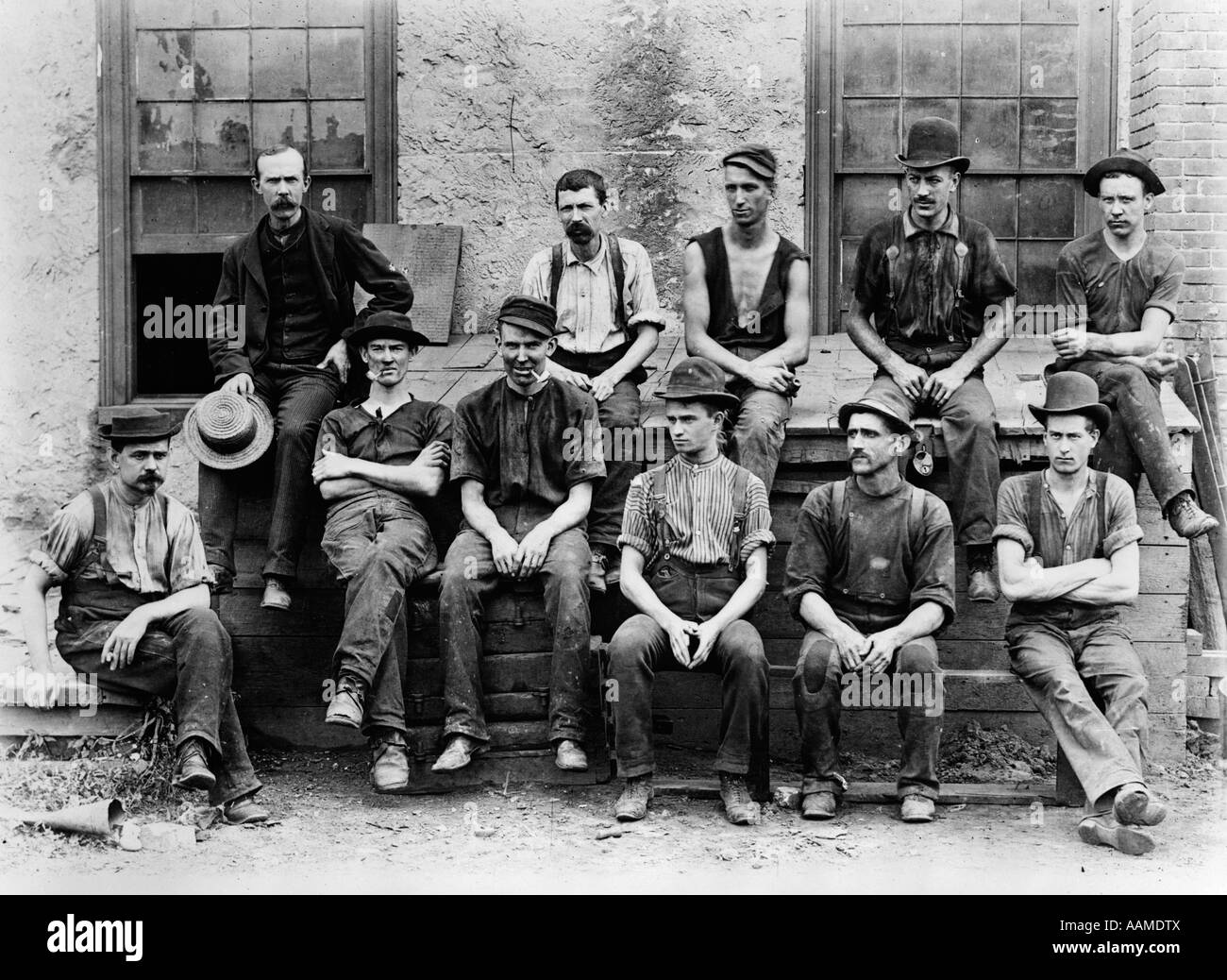 1890s 1900s GROUP OF 11 FACTORY WORKERS SEATED OUTSIDE OF BUILDING Stock Photo