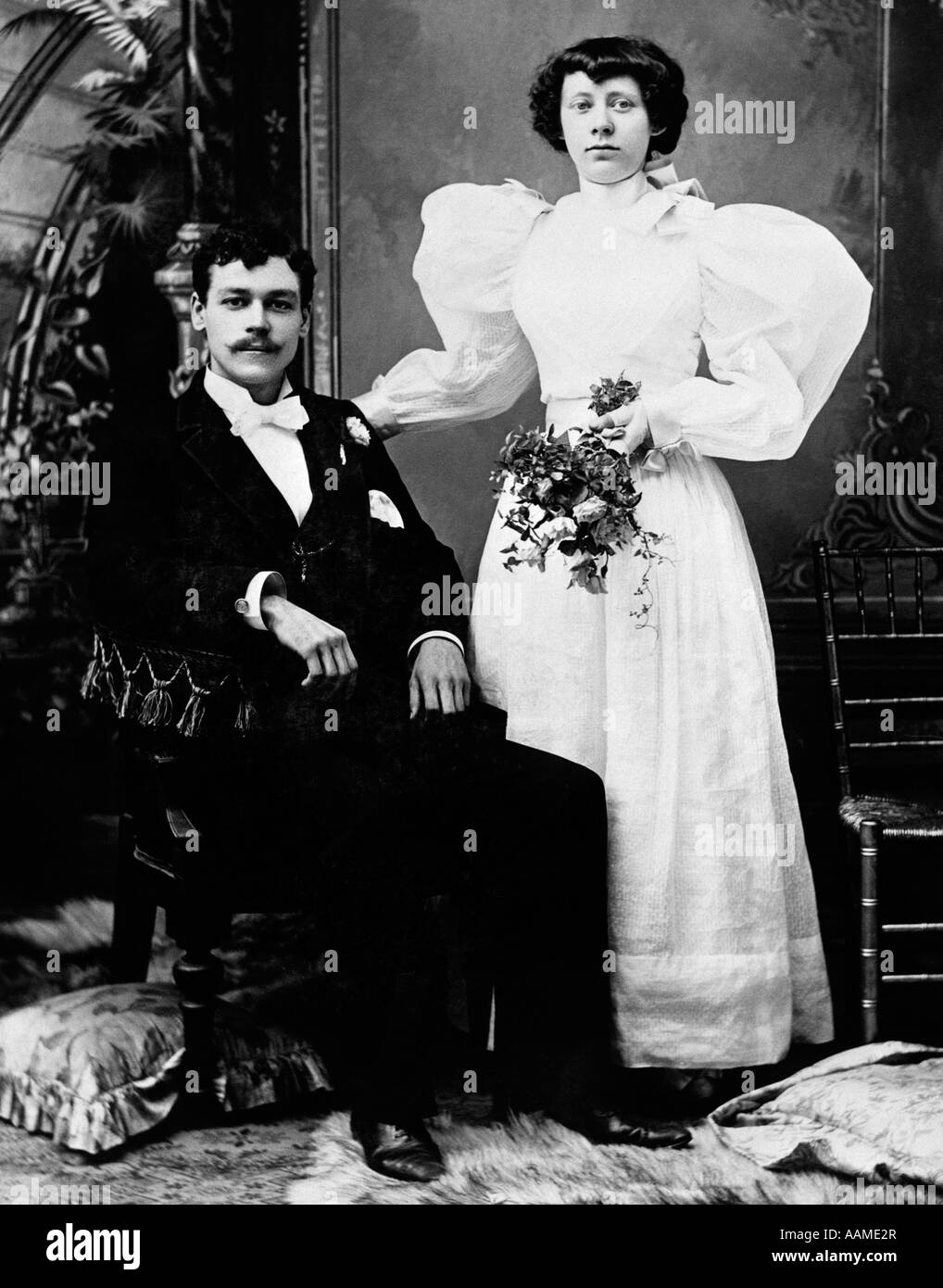 EARLY 1900s FORMAL PORTRAIT OF BRIDE AND GROOM MAN SEATED WOMAN STANDING INDOOR Stock Photo