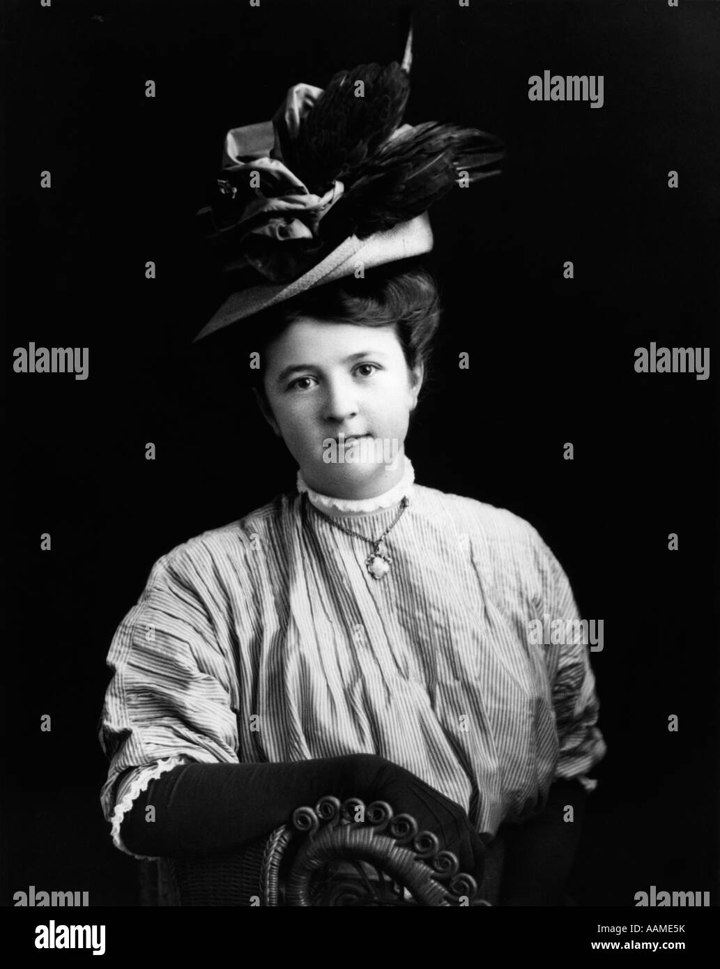 1890s 1900s TURN OF THE CENTURY PORTRAIT WOMAN SEATED IN WICKER CHAIR WEARING FANCY HAT & LONG GLOVES Stock Photo