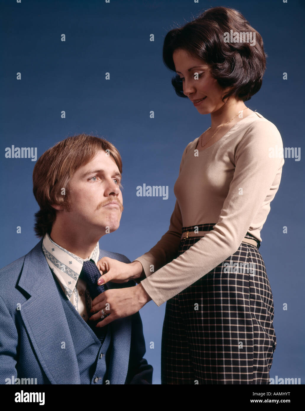 1970s COUPLE WOMAN FIXING TIE NECKTIE OF MAN SEATED BESIDE HER HUSBAND WIFE FASHION RETRO VINTAGE BLUE BACKGROUND Stock Photo