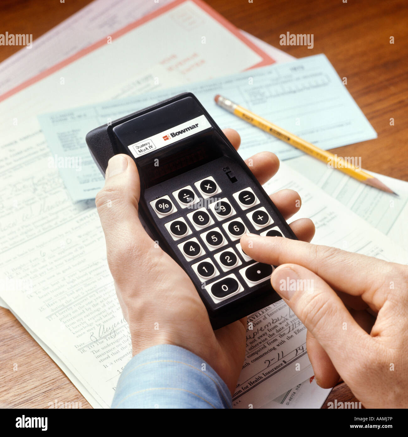 1970s DETAIL OF HANDS HOLD CALCULATOR NUMBERS INDEX FINGER ABOUT TO PUSH BACKGROUND OF BILLS PAPERS ON TABLE Stock Photo