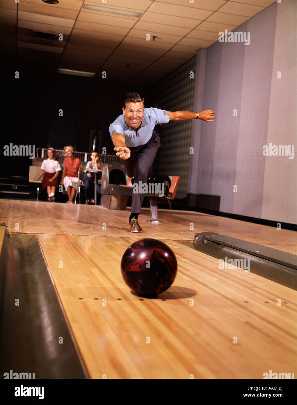 1960s 1970s SMILING MAN THROWING BOWLING BALL DOWN ALLEY AS FAMILY WATCHES IN BACKGROUND FUN DAD FATHER RETRO VINTAGE Stock Photo