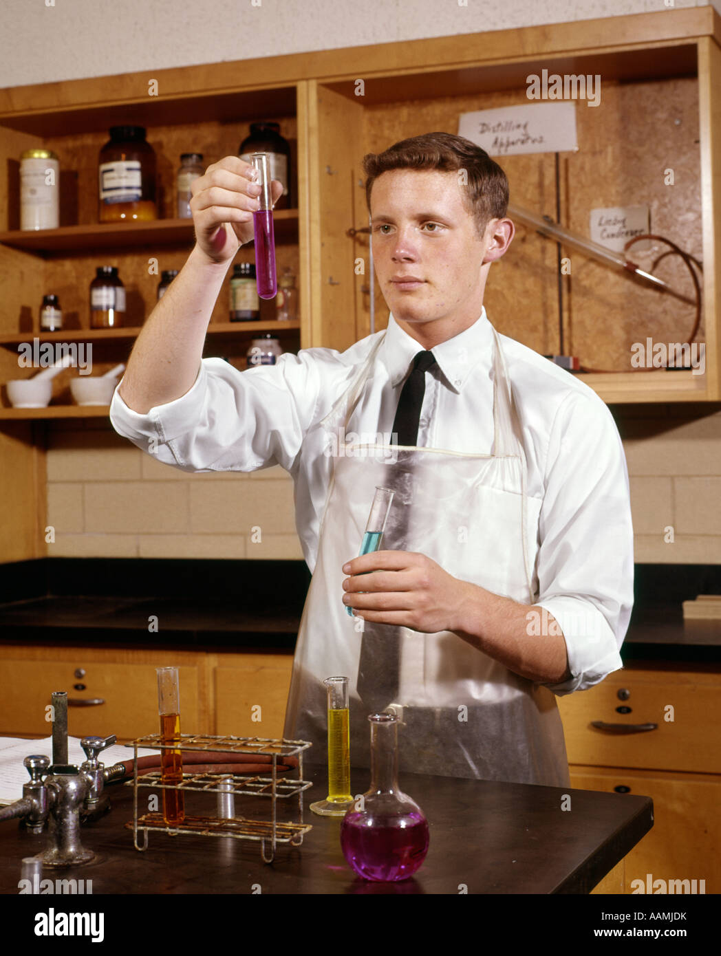 1960s MAN COLLEGE STUDENT IN CHEMISTRY LABORATORY LOOKING AT SOLUTION IN VIAL EXPERIMENT LEARN ONE OBSERVE TEST Stock Photo
