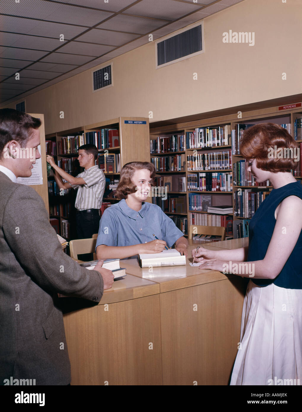 1960s FOUR STUDENTS IN COLLEGE LIBRARY THREE AT TABLE AND ONE IN STACKS BOOK SHELF STUDY EXAMINATION LESSONS Stock Photo