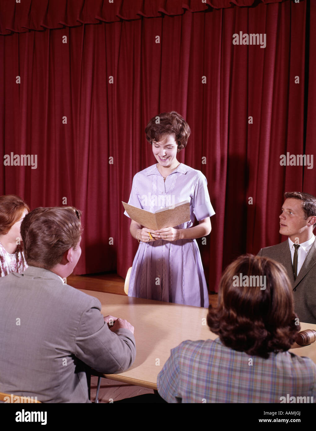 1960s TEEN STUDENTS AT CONFERENCE TABLE IN FRONT OF RED VELVET CURTAIN GIRL OPEN FOLDER SPEAK STUDENT GOVERNMENT COUNCIL Stock Photo
