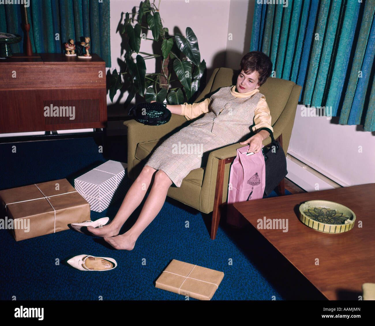 1960s WOMAN TIRED EXHAUSTED IN LIVING ROOM CHAIR SORE FEET SHOES OFF SHOPPING BAGS ALL AROUND